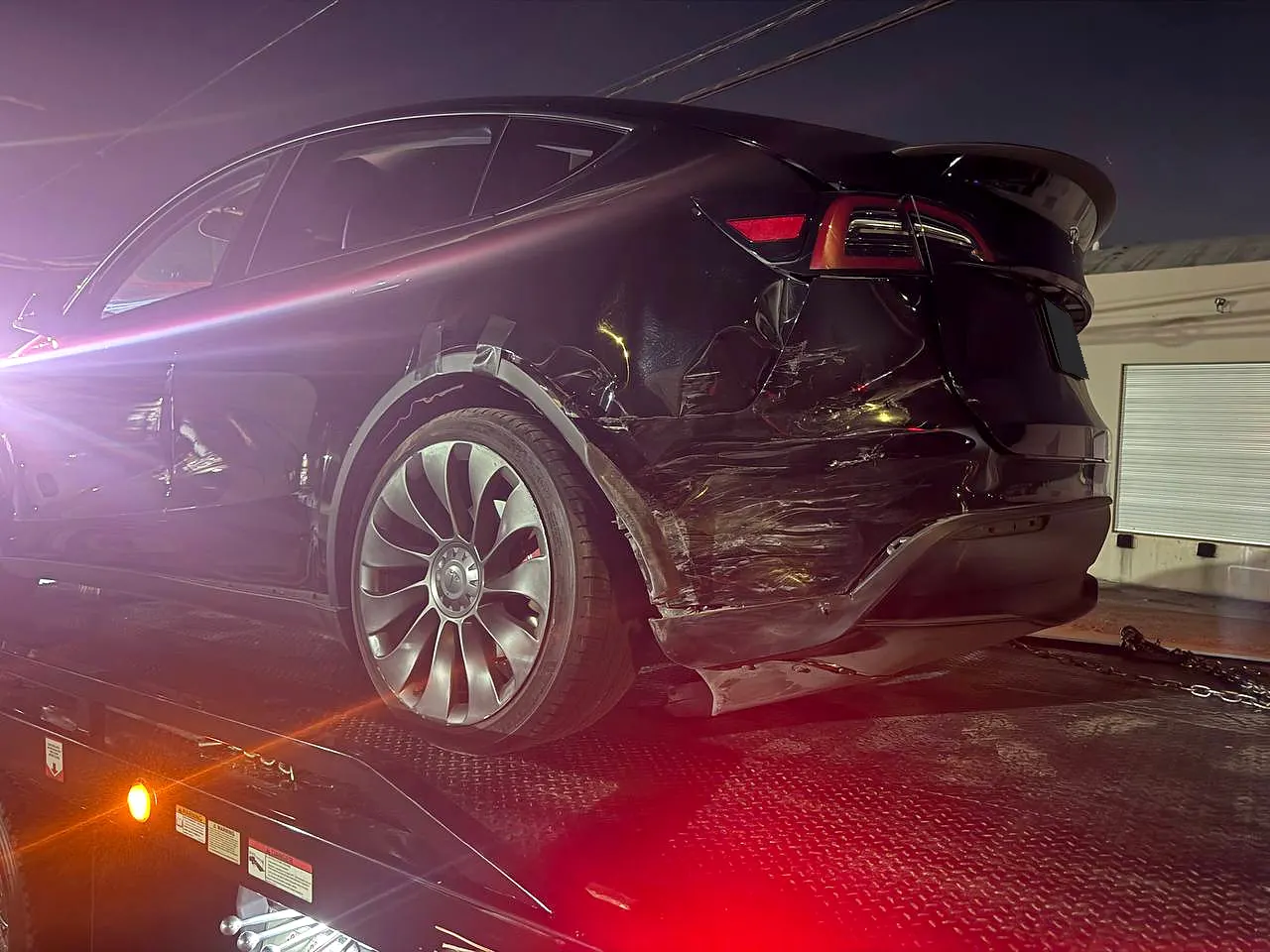 Tesla with visible rear collision damage on a flatbed tow truck at night
