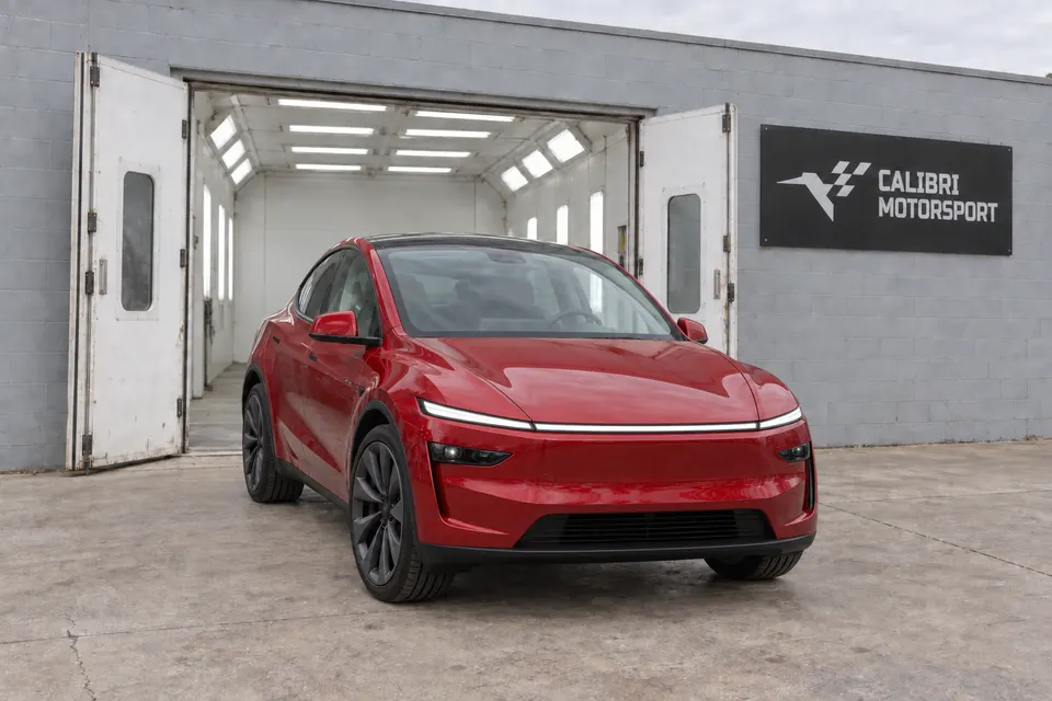 Red refreshed Tesla Model Y outside a paint booth built into the gray shop facade, with Calibri Motorsport signage